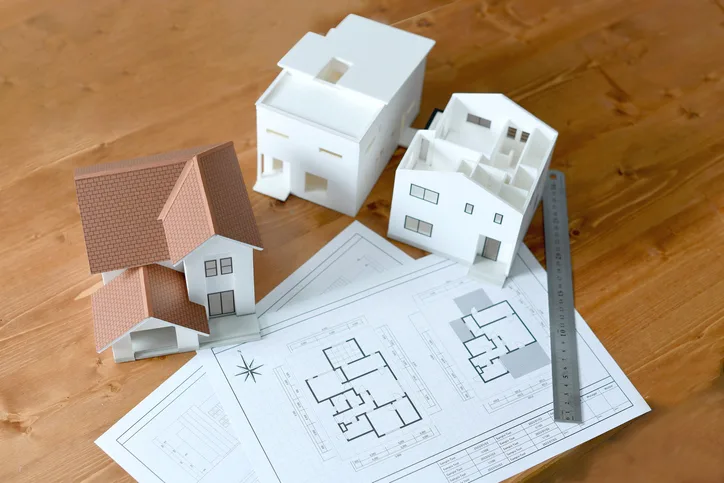 Weiße Architekturmodelle von Einfamilienhäusern neben einem Grundriss-Bauplan und einem Lineal auf einem Holztisch